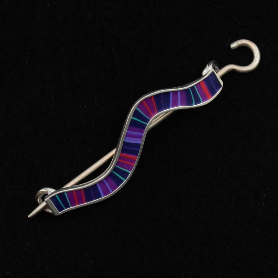 The Stripe Wavy Shawl Pin by Bonnie Bishoff Designs features a beautiful, curved silver design with a colorful geometric pattern in shades of purple, blue, and red. Handmade by the talented artisans at Bonnie Bishoff, this brooch includes a hook on one end and a pin clasp on the other, set against a black background—an ideal accessory for knitted wearables.