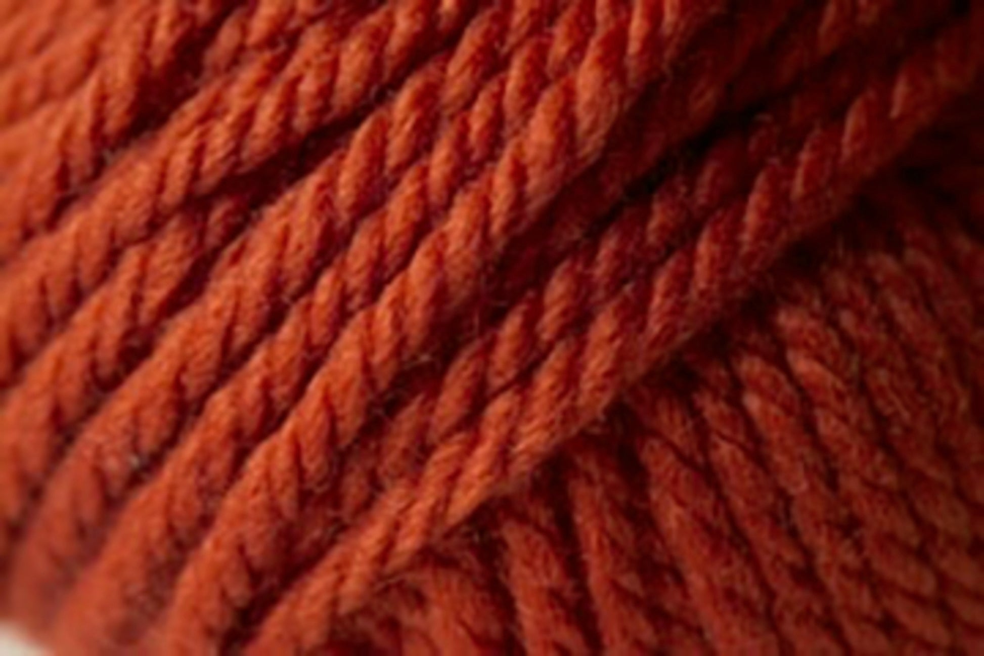 Close-up image of thick, rust-colored Jo Sharp Silkroad Ultra yarn by Kingfisher Yarn & Fibre. The twisted fibers form a tightly wound coil, displaying a rich and earthy tone. The texture of the chunky cuddly yarn appears super soft and suitable for knitting or crocheting cozy winter accessories.