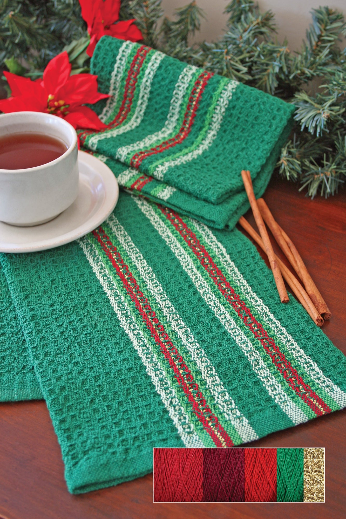 A white cup filled with tea sits on green cloth napkins adorned with red and white stripes. Next to the cup are several sticks of cinnamon. In the background, there is festive greenery and red poinsettias. The bottom right corner features the Waffle Weave Dish Towel Kit from Halcyon Yarn, showcasing vibrant patterns in red, green, and gold yarns.