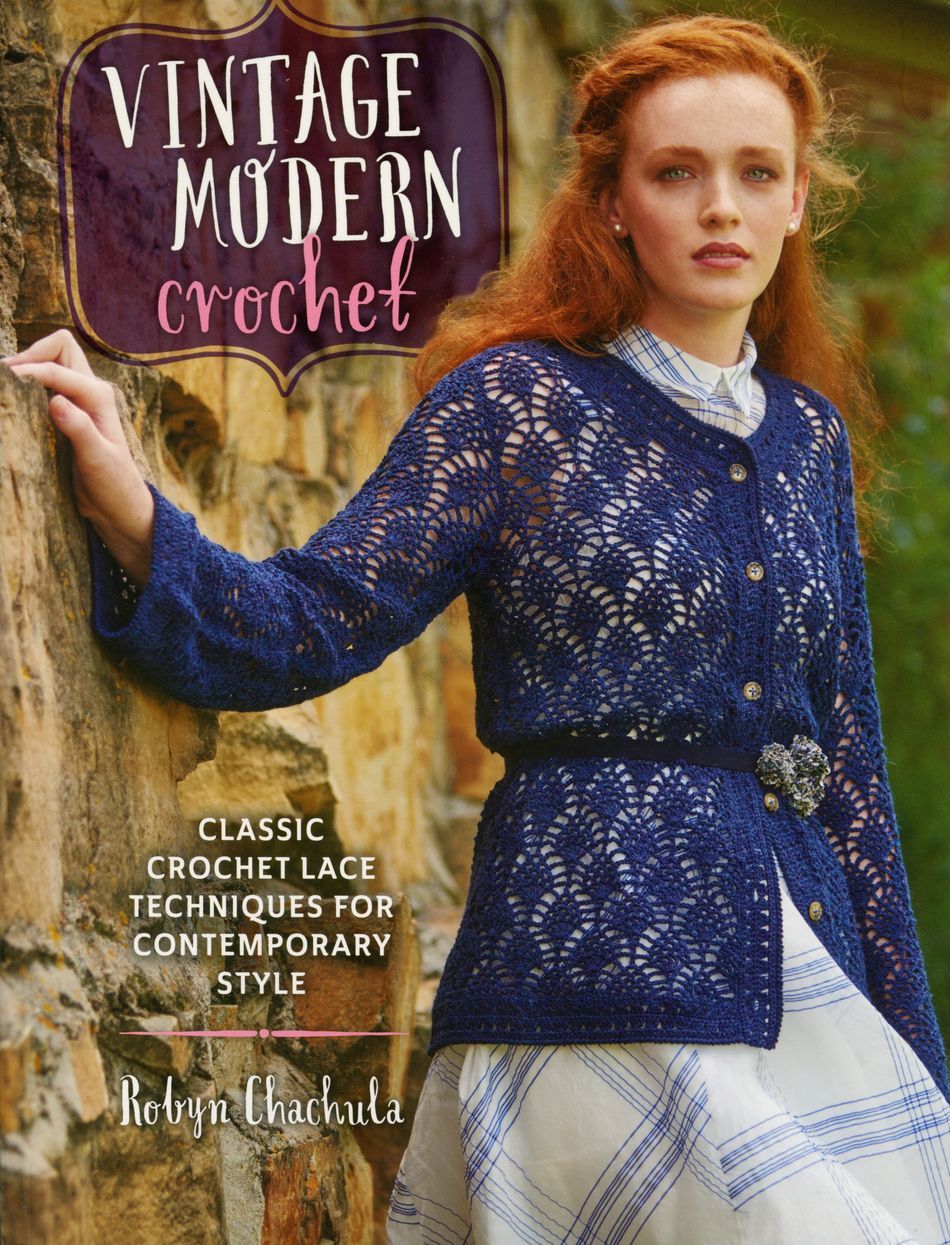 A woman with long, wavy red hair leans against a stone wall, adorned in a dark blue lace crochet cardigan over a white dress with blue stripes. The book titled "Vintage Modern Crochet" by Ingram Content highlights classic crochet lace techniques.