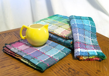 A yellow ceramic pitcher is placed on a wooden table next to a stack of colorful kitchen towels from the Halcyon Yarn's Homestead Cotton Color Gamp Kit. The towels, resembling a successful weaving project with their various plaid patterns and vibrant hues, add a cozy and cheerful aesthetic to the scene. A white curtain is in the background.