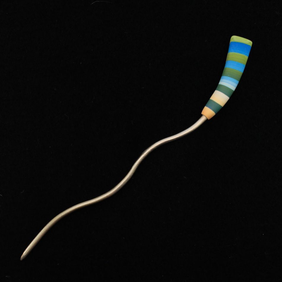 The Striped Color Stick Shawl Pin by Bonnie Bishoff Designs, from the Bonnie Bishoff brand, features a wavy metallic tip and a colorful polymer clay handle with green, blue, yellow, and white stripes, set against a black background. The metal design curves smoothly for an elegant finish.