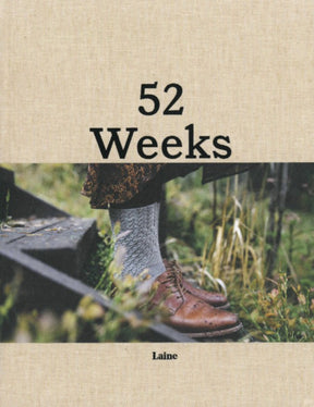 A book cover titled "52 Weeks of Socks" by Laine USA Distribution. This beige coffee table book showcases a close-up of a person wearing textured, hand-knitted gray socks paired with brown leather shoes, set against a background of tall grass. The partial view of wooden outdoor steps enhances its Nordic-style knitting charm.
