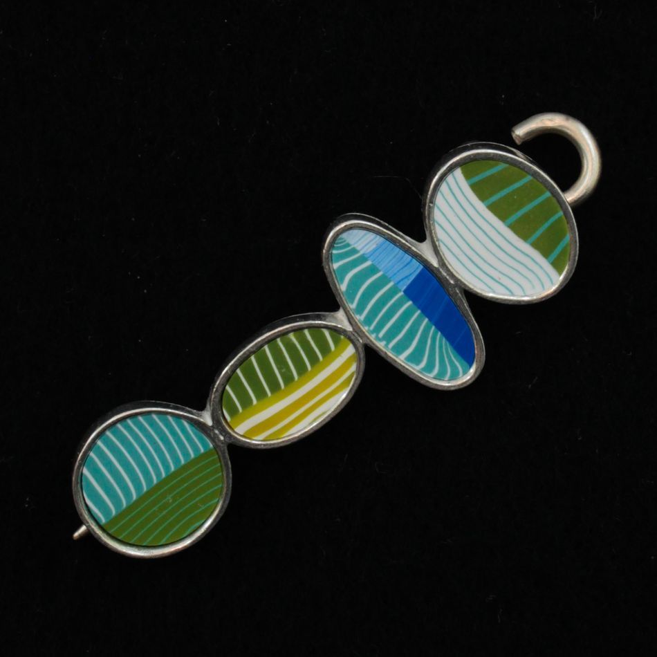 The Bubbles Shawl Pin by Bonnie Bishoff Designs from the brand Bonnie Bishoff features four linked, oval-shaped elements with colorful striped designs. The ovals, bordered in silver, showcase teal, green, blue, and yellow stripes on a black background reminiscent of intricate polymer clay design. The piece includes a pin on the back and a hook on one end for secure fastening.