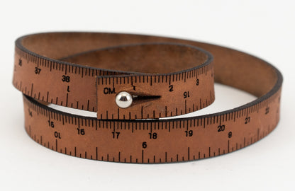 A close-up image of the Leather Wrist Ruler by Bryson Distributing, Inc. showcases a medium brown leather wrap-around bracelet designed to resemble a measuring tape. The wristband features black measurement markings and numbers in centimeters and is coiled in a circular shape with a metal stud closure.