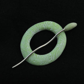 The Carved Ring Shawl Pin by Bonnie Bishoff Designs, crafted by Bonnie Bishoff, is a circular green piece with an intricate white pattern. Ideal for securing lightweight shawls or adorning knit wearables, it stands out against a black background. A textured metallic pin runs through its center, adding to the elegant and detailed design.