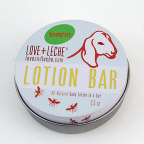 A round tin container of Love + Leche Lotion Bar, featuring a goat illustration and text that reads "All Natural body lotion in a bar, 2.5 oz," is perfect for keeping skin deeply moisturized. Infused with beeswax, the white and silver container is adorned with red and yellow text, plus small images of bees.