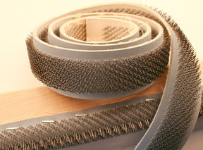 A close-up image of a coiled Rug Hooking Gripper Strip by Stratosphere Inc - Howard Brush Co on a flat surface. The strip is wrapped around a piece of wood, resembling a quilter's pin setup, with additional lengths of the strip with parallel rows of tiny, sharp pins visible in the background.