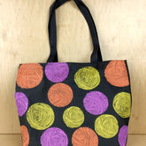 The Polka Dot Yarn Tote by Bonnie Bishoff is a black Cotton-Poly Canvas tote bag featuring a vibrant design of yarn balls in orange, yellow, and purple against a plain wooden background. It comes with two black handles and pairs perfectly with the Bonnie Bishoff shawl pins.