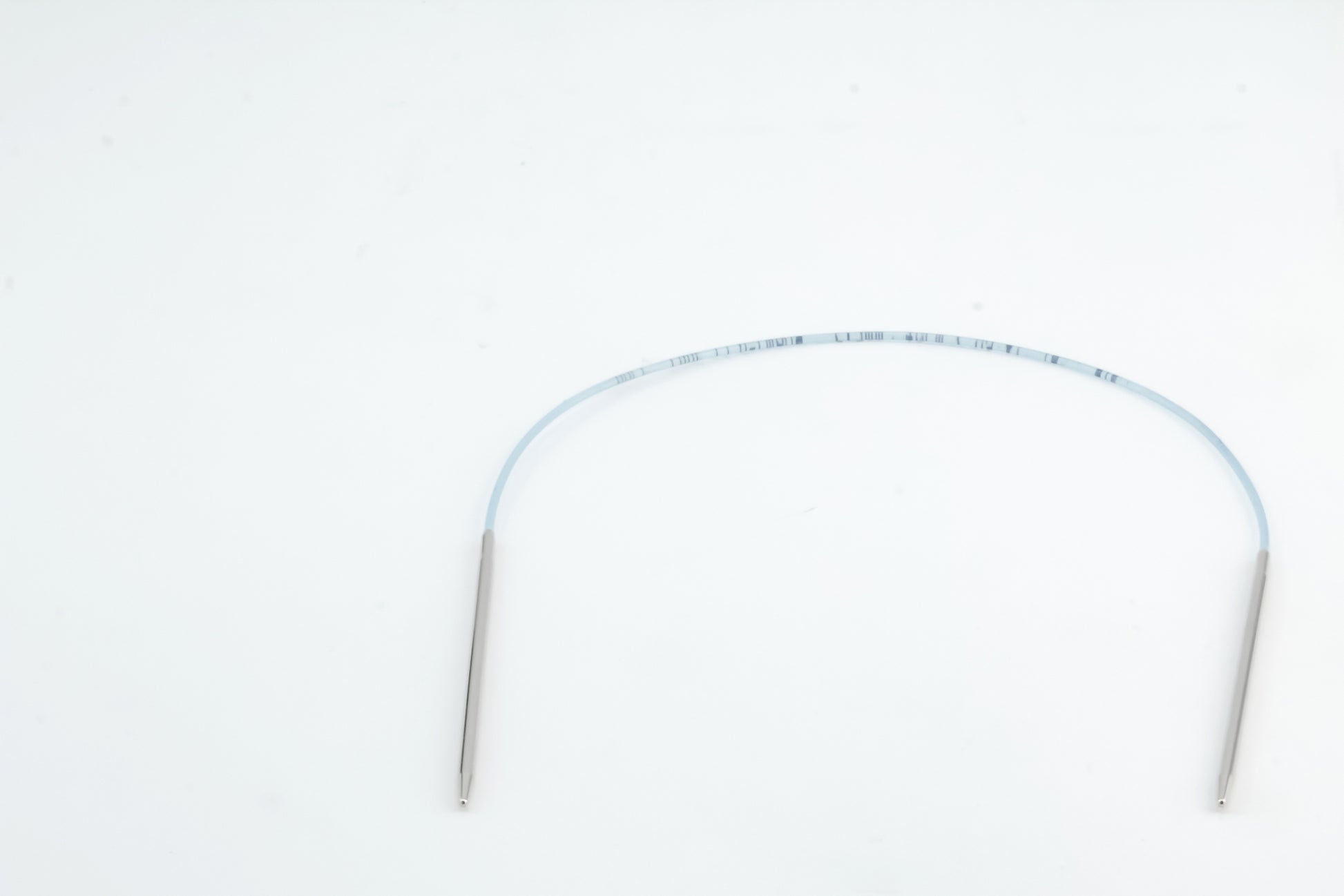 An image depicting a thin, flexible, translucent medical catheter with two metallic ends resting on a plain white surface. The catheter is gently curved and features markings along its length, evoking the precision of Skacel's Addi Turbo Circular Knitting Needles used for crafting intricate doll clothes.