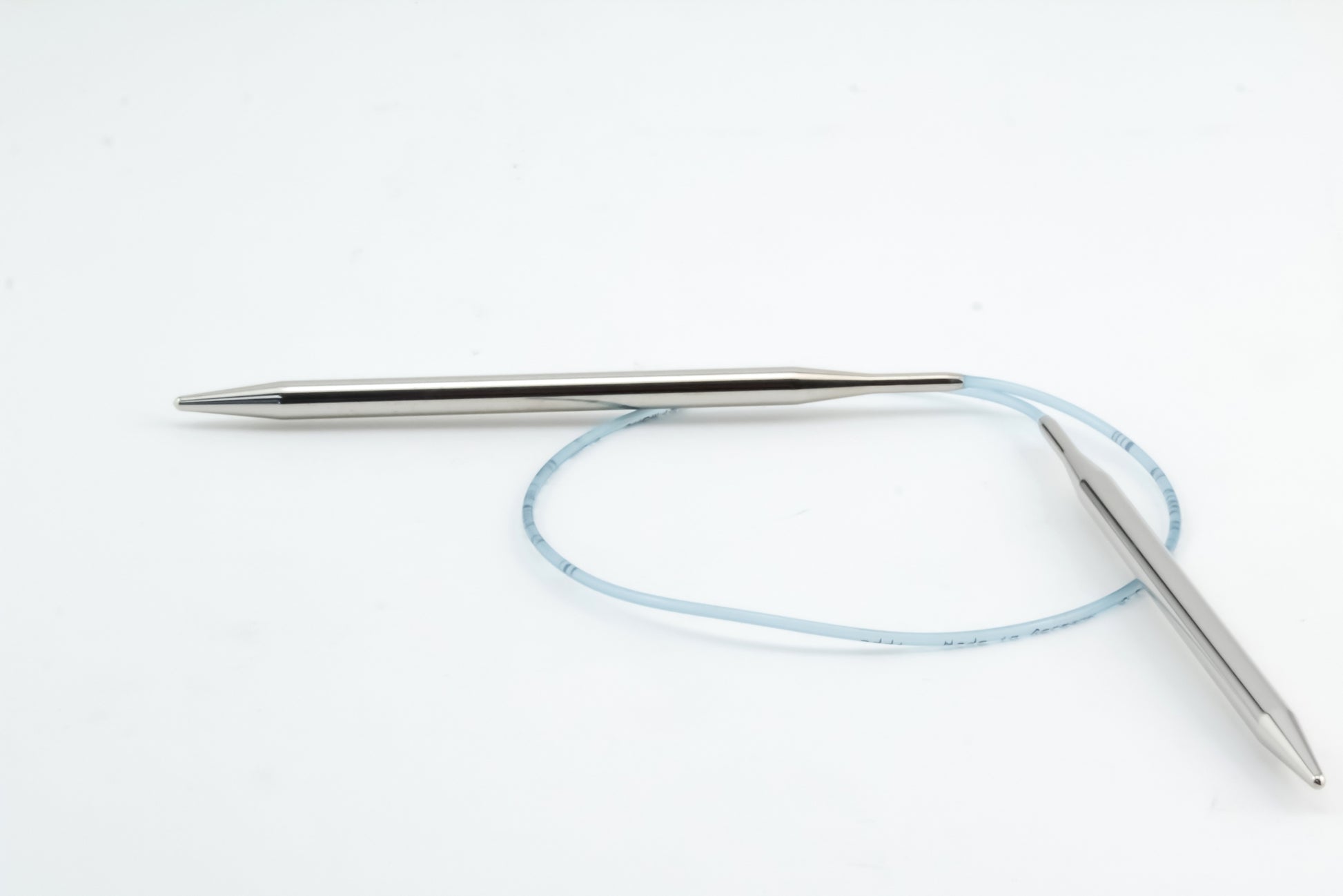 A pair of Skacel Addi Turbo Circular Knitting Needles, characterized by their metal construction and connected by a flexible blue cable, is placed on a white background. One of the needles is positioned horizontally while the other lies diagonally, with the cable partially looped between them. These needles are ideal for crafting cuffs, collars, and doll clothes.