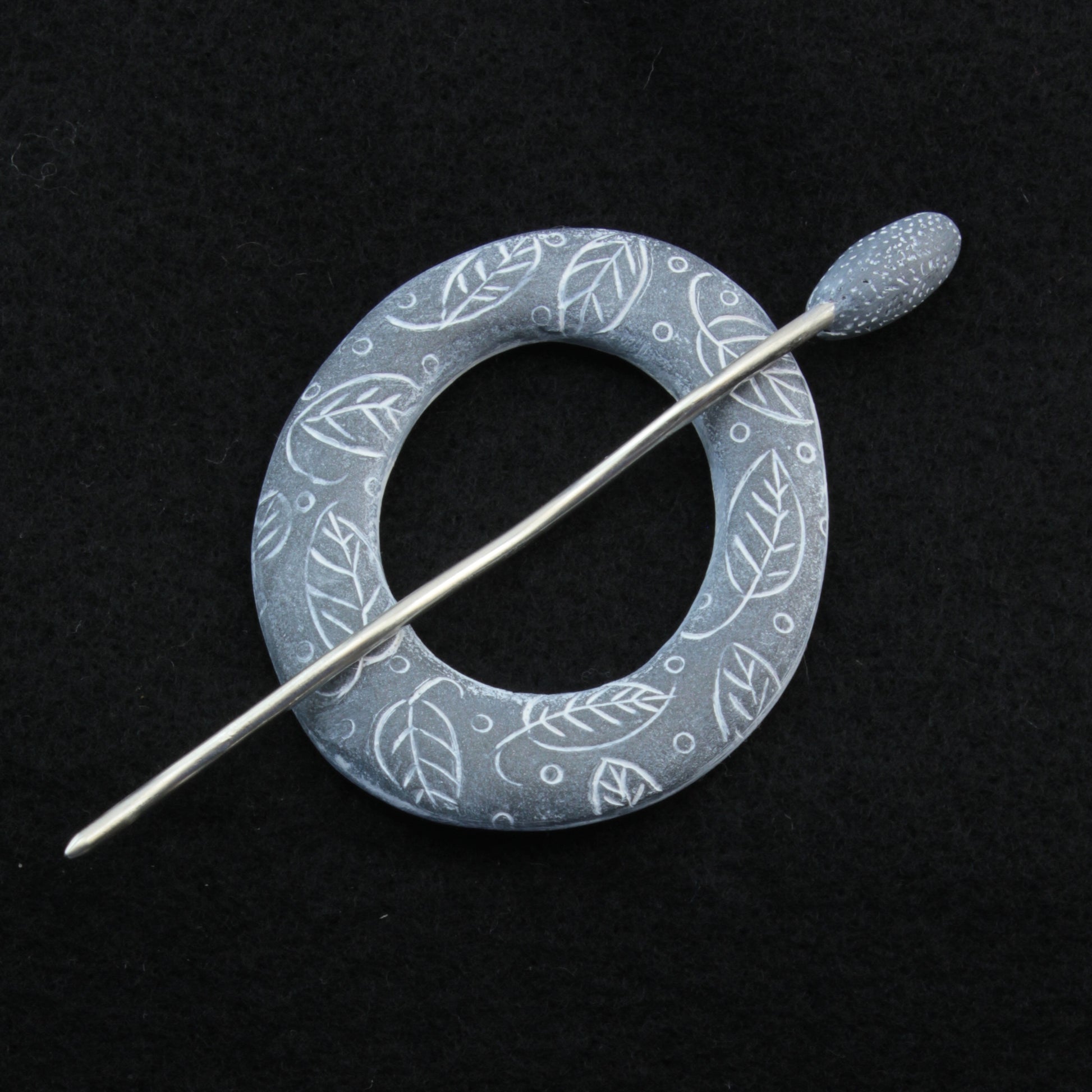 The Carved Ring Shawl Pin by Bonnie Bishoff Designs features intricate white leaf patterns on a round brooch in shades of blue, complemented by a silver stick pin for an elegant, nature-inspired look. Perfect as a lightweight shawl closure, the black background beautifully highlights the polymer clay design details of this exquisite piece from Bonnie Bishoff.