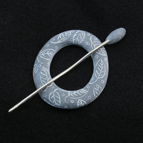 The Carved Ring Shawl Pin by Bonnie Bishoff Designs features intricate white leaf patterns on a round brooch in shades of blue, complemented by a silver stick pin for an elegant, nature-inspired look. Perfect as a lightweight shawl closure, the black background beautifully highlights the polymer clay design details of this exquisite piece from Bonnie Bishoff.