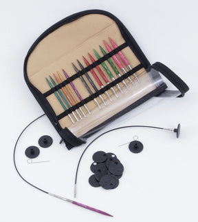 An Accessories Unlimited Dreamz Interchangeable Circular Knitting Needle Set in a tan and black foldable case is open, displaying colorful densified laminated birch needle tips in various sizes. The set includes black circular cables, caps, stoppers, and tightening tools, arranged neatly beside the case.