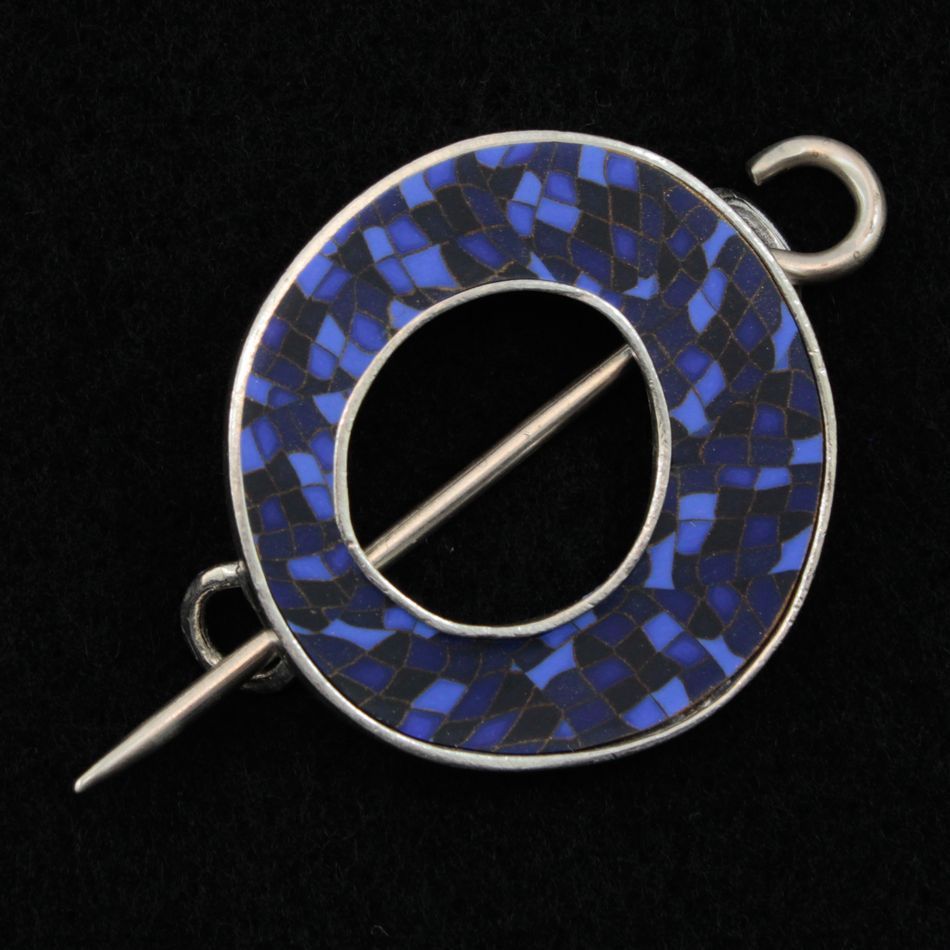 The Single Circle Shawl Pin by Bonnie Bishoff Designs displays a stunning blue and black mosaic pattern, meticulously crafted from polymer clay. Featuring a central hole and a metal pin running across the middle, this piece showcases small square tiles arranged in a woven appearance. The pin's round loops at both ends make it an ideal accessory for knit wearables, brought to you by the renowned brand Bonnie Bishoff.