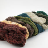 A pile of multicolored wool roving, ideal for felting projects, lies on a white background. The Harrisville Fiber Batts Collection by Halcyon Yarn features various shades, including deep red, brown, yellow, olive green, forest green, and teal. The fibers are loose and fluffy, ready for spinning or crafting multi-color bags.