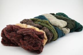 A pile of multicolored wool roving, ideal for felting projects, lies on a white background. The Harrisville Fiber Batts Collection by Halcyon Yarn features various shades, including deep red, brown, yellow, olive green, forest green, and teal. The fibers are loose and fluffy, ready for spinning or crafting multi-color bags.