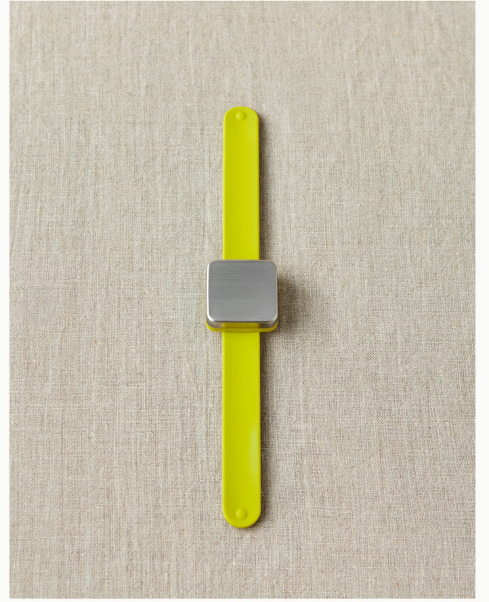 A minimalist Maker's Keep Magnetic Bracelet from Cocoknits, featuring a silver square face and a bright yellow silicone slap bracelet, is placed on a textured beige surface. The bracelet is designed with a sleek, modern look, and the band appears to be made of flexible material.