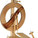 The Kromski Fantasia Spinning Wheel by Kromski North America is a wooden spinning wheel with a modern double drive wheel design. It features a large circular wheel, two foot pedals, and various smaller components, including Kromski bobbins and hooks. The spinning wheel boasts a sleek natural wood finish with darker wood accents.