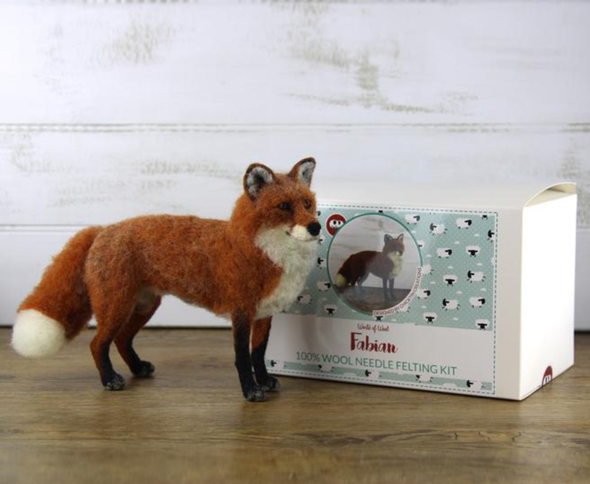 A needle-felted fox figurine stands beside a box labeled "Animal Sculpture Felting Kits" by Europa Wools Ltd. The packaging, which features the realistic animal and text that reads "Fabian," highlights the intricate use of wool fibers. The background is a rustic white wooden surface.