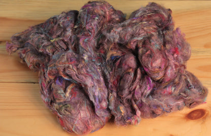 A quarter-pound bag of Indian Silk Shop's Pulled Sari Silk Fiber, featuring a medley of multicolored and textured wool roving, rests on a wooden surface. This fluffy, fibrous blend is interspersed with vibrant hues of purple, pink, red, and blue.