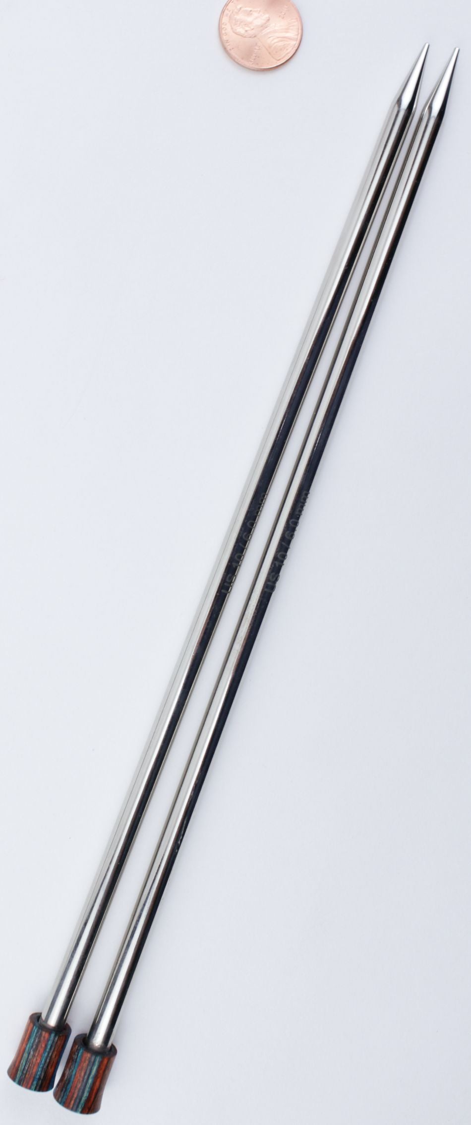 Two long, shiny metallic Nova Platina Single Point Knitting Needles from Accessories Unlimited with tapered tips, arranged parallel to each other on a white background. A penny is placed above the needles for size comparison. The needles have maroon-colored ends and resemble sleek hollow brass pipes.