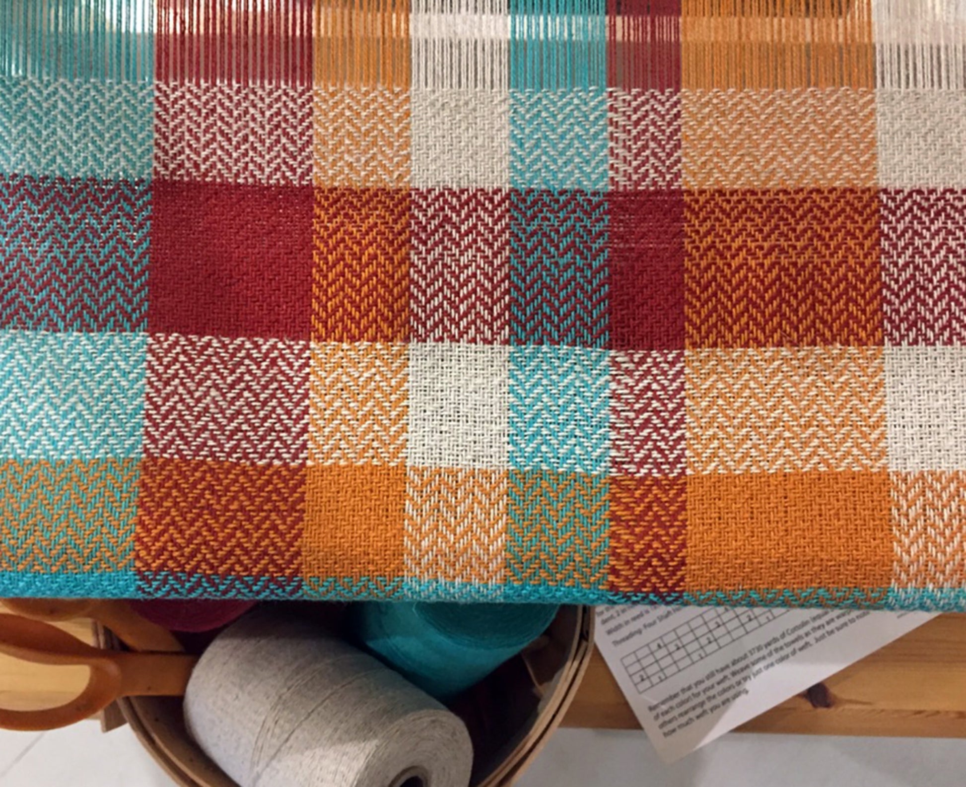 A close-up view of a woven fabric showcasing a symmetrical plaid pattern from the Organic Cottolin Tea Towel Kit by Halcyon Yarn. The design includes vibrant colors such as red, orange, blue, and white. Below the fabric, spools of thread and a piece of paper with text are partially visible.