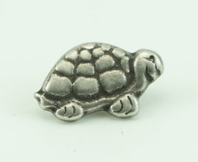 A small, silver-toned metal sculpture of a turtle with a detailed shell and limbs, reminiscent of the intricate design found on an 11/16" diameter Pewterette Button by Buttons Etc. The turtle's head and legs feature simple, carved markings, and it is displayed against a plain white background.