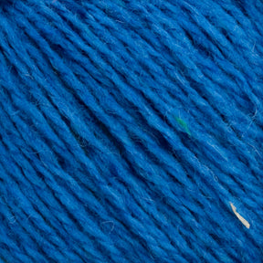 Close-up view of a section of unscoured yarn from the Harrisville Designs Harrisville Highland - Cones, showcasing the texture and individual threads. The yarn appears soft and slightly fuzzy, with a few small fibers and spinning oils visible. Subtle variations in the blue coloration and a few small specks of other colors add character.