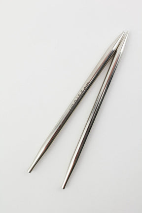 A pair of sleek, metallic knitting needles from Accessories Unlimited's Nova Platina Interchangeable Knitting Needles collection lies side by side on a plain white surface. The 4.5" tips are sharply pointed at one end and feature a small engraved branding near the top. Their smooth, polished surface reflects light subtly.