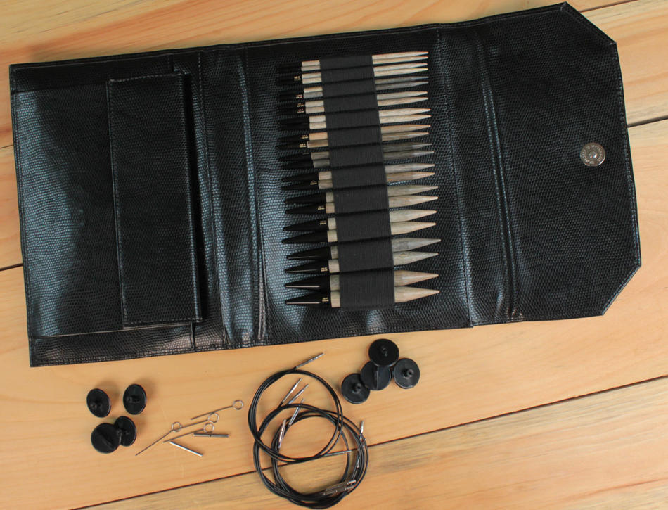 A black leather case containing a set of Knitting Fever / Euro Yarns Lykke Interchangeable Circular Knitting Needles, 5" with black caps is open on a wooden surface. Beside the case are circular cables, black plastic buttons, and several metal accessories like a cable key and stoppers from the Lykke needle set.