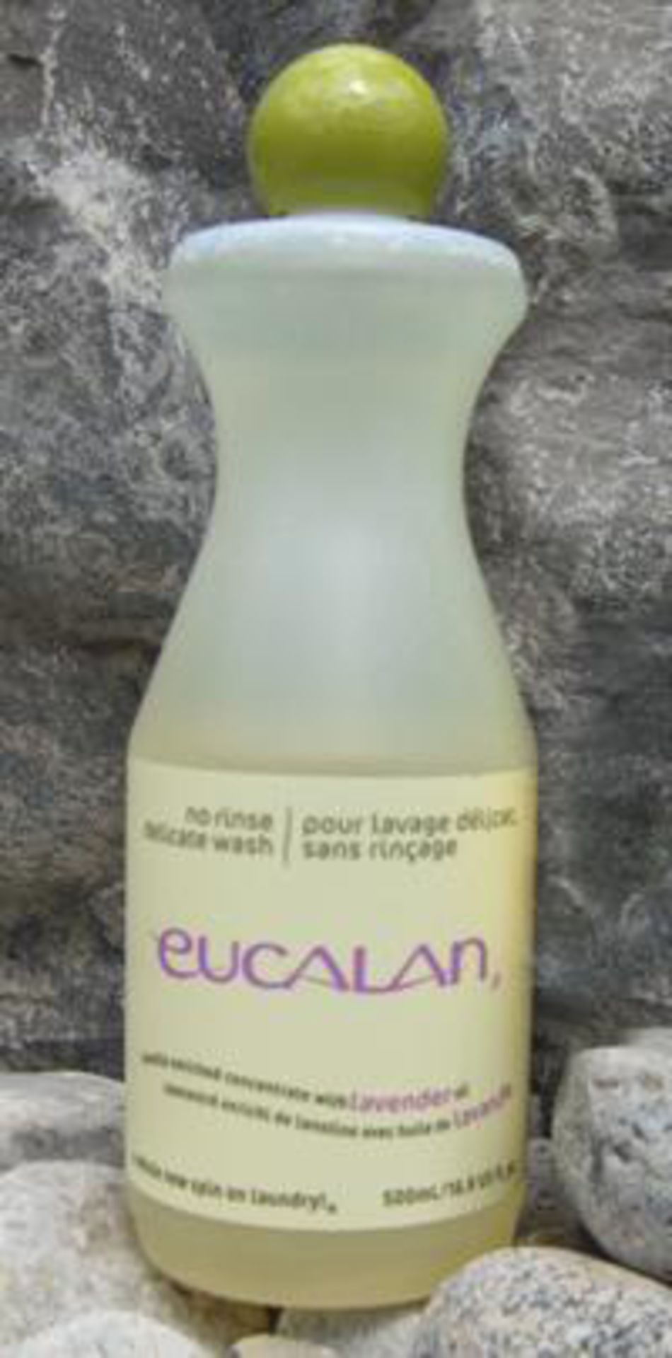 A 16.9 oz bottle of "Eucalan Wool Wash" with a lavender scent is placed against a stone background. This lanolin-enriched concentrate is translucent white with a green cap and purple text. Labeled in both English and French, it boasts a 500ml capacity and is 100% biodegradable.