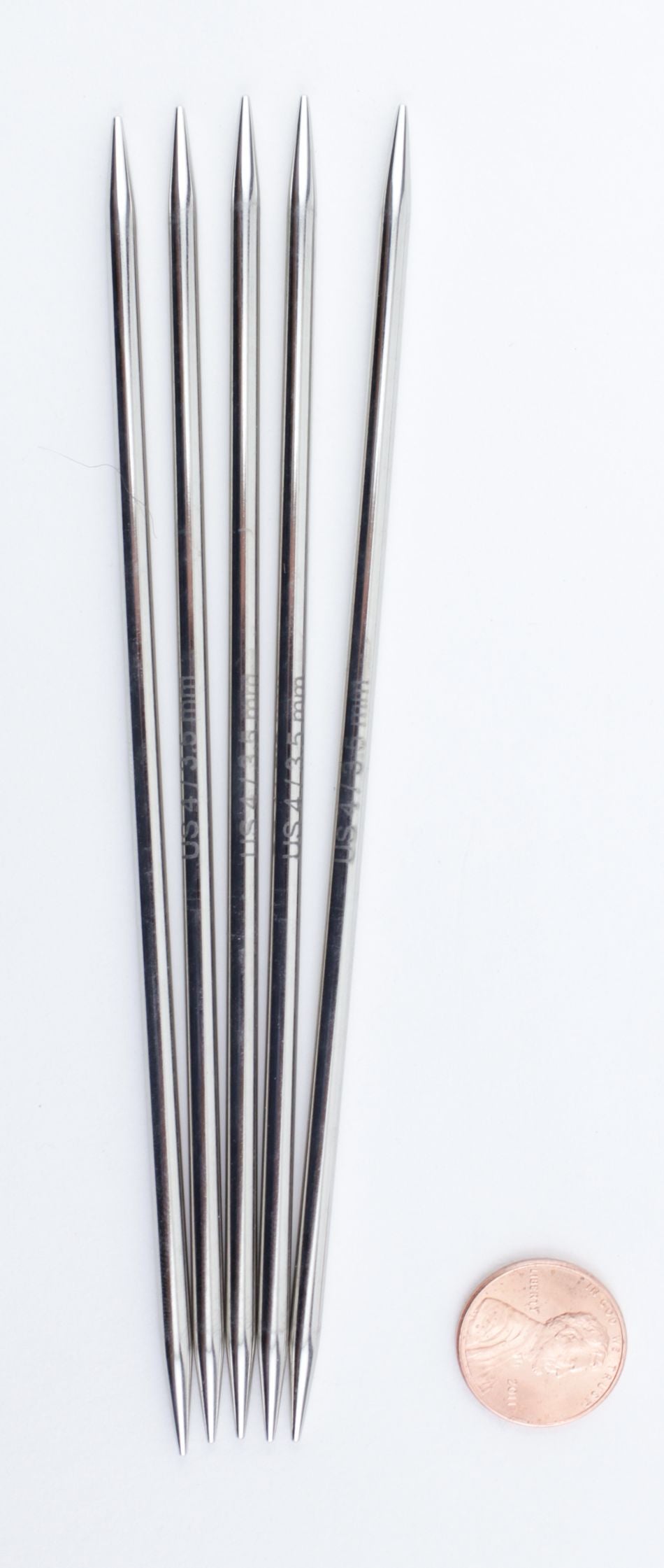 A set of five Nova Platina Double-Point Knitting Needles from Accessories Unlimited is lined up side by side with their pointed ends facing up. To the right of these high-quality chrome needles is a U.S. penny, providing a sense of scale. The reflective surface of these knitting needles shines brilliantly against a white background.