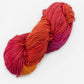 A skein of Great Adirondack Organic DK Cotton by Great Adirondack Yarn Co, Inc., hand-dyed and multi-colored with hues ranging from deep red to vibrant orange and magenta, lying against a white background.