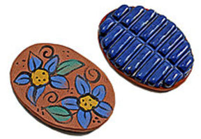 Two oval clay tokens from the Felting Stone collection by Kick the Moon Farm: one showcases hand-painted blue flowers with green leaves and yellow centers on a terracotta background; the other boasts a textured, raised blue pattern reminiscent of waves or tiles on a red-brown background, evoking the intricate designs seen in traditional felting.