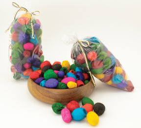 Three transparent bags filled with vibrant Dyed Silk Cocoons from Indian Silk Shop, tied with twine bows, are displayed beside a wooden bowl also containing these colorful cocoons. The hues of the cocoons stand out beautifully, resembling shades seen in dyed silk: red, blue, yellow, green, and purple.