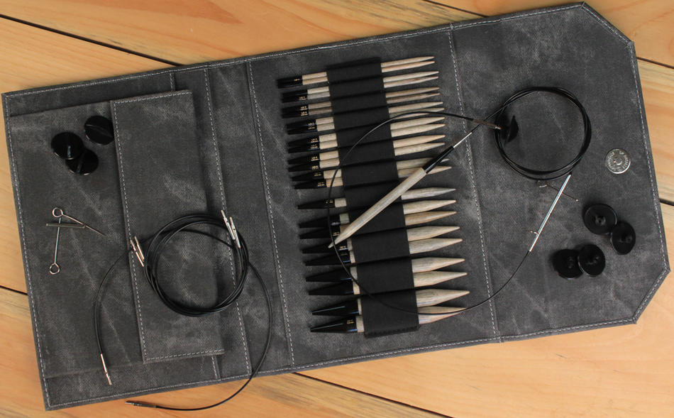 The Lykke Interchangeable Circular Knitting Needle Set, 5" by Knitting Fever / Euro Yarns is neatly organized in a gray fabric case. The set includes multiple pairs of interchangeable birch knitting needles of various sizes, black cables, black caps, and other knitting accessories. Everything is arranged meticulously on a light wooden surface.