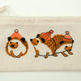 Introducing the Wee Hedgehog Zip Pouch by Mum n Sun Ink, crafted by Bonnie Bishoff. This cream-colored zippered pouch is made from Envirotote canvas and features three charming hedgehog illustrations. The left hedgehog is curled into a ball, the middle one is standing, and the right one is walking away—all adorned with red and white winter hats. Perfect for storing shawl pins or small essentials.