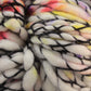 Close-up of a skein of thick, multi-colored superwash merino wool from Malabrigo Yarn. The Malabrigo Caracol is primarily white with interwoven black strands and features splashes of yellow, purple, and red hues. This kettle-dyed super bulky yarn has a soft, fuzzy texture.