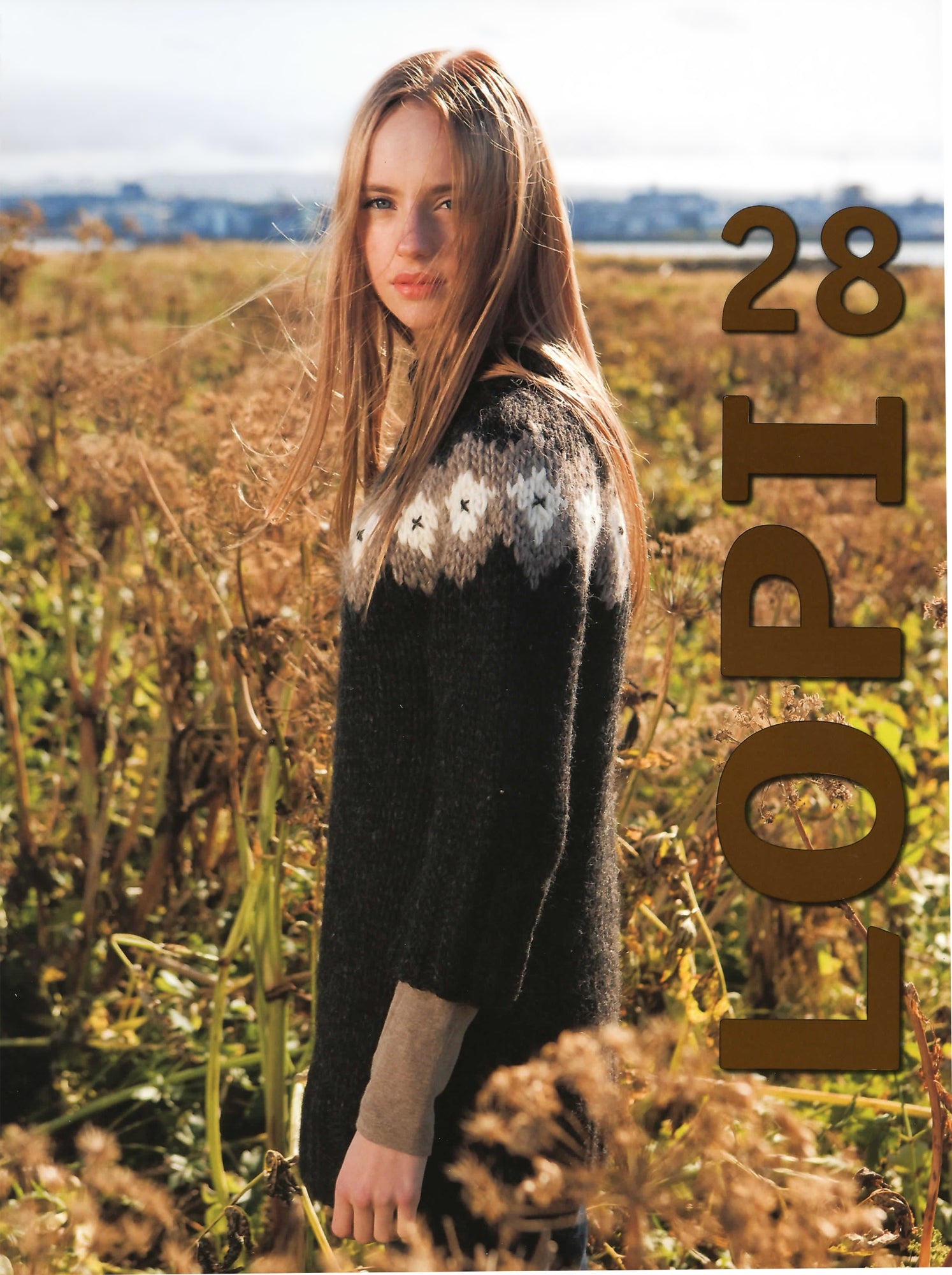 A woman stands in a field of dried vegetation, donning a black and white patterned sweater. The text "Lopi Volume 28" is prominently displayed on the right side of the image. In the background, a distant view of a water body and some buildings can be seen. This beautiful piece is part of Berroco, Inc.'s stunning collection.