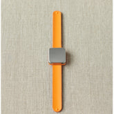 The Maker's Keep Magnetic Bracelet by Cocoknits, featuring an orange band and a square silver face, is displayed on a light gray fabric background. Resembling the innovative style of a silicone slap bracelet, the watch is centered and positioned vertically in the image.