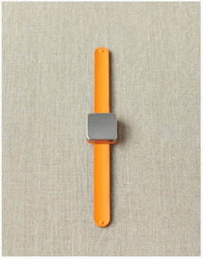 The Maker's Keep Magnetic Bracelet by Cocoknits, featuring an orange band and a square silver face, is displayed on a light gray fabric background. Resembling the innovative style of a silicone slap bracelet, the watch is centered and positioned vertically in the image.