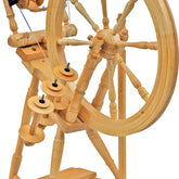 The Kromski Interlude Spinning Wheel by Kromski North America, featuring a single drive angle treadle design, boasts a large wheel on the right side connected to several smaller spindles and components on the left. Crafted from European alder and birch, its frame showcases intricate woodworking details.