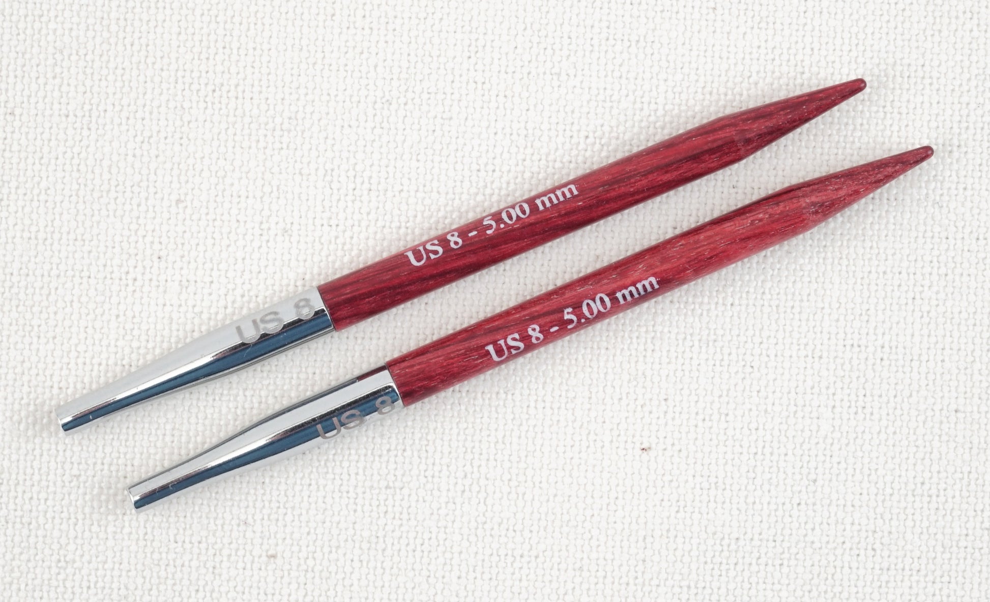 Two Dreamz Interchangeable Knitting Needle Tips, size US 8 (5.00 mm), from Accessories Unlimited are laid parallel on a white fabric surface. The needle tips are made of metal, with the body of each needle crafted from wood stained in a red hue, featuring smooth joins perfect for knitting hats effortlessly.