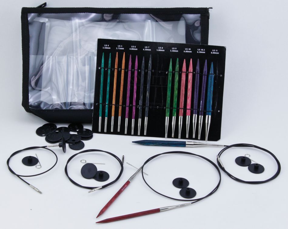 The Dreamz Interchangeable Circular Knitting Needle Set by Accessories Unlimited, showcasing a variety of sizes in vibrant patterns and made from densified laminated birch, is elegantly displayed on a white surface next to black cables and needle accessories. A clear zippered carrying case is partially visible in the background.