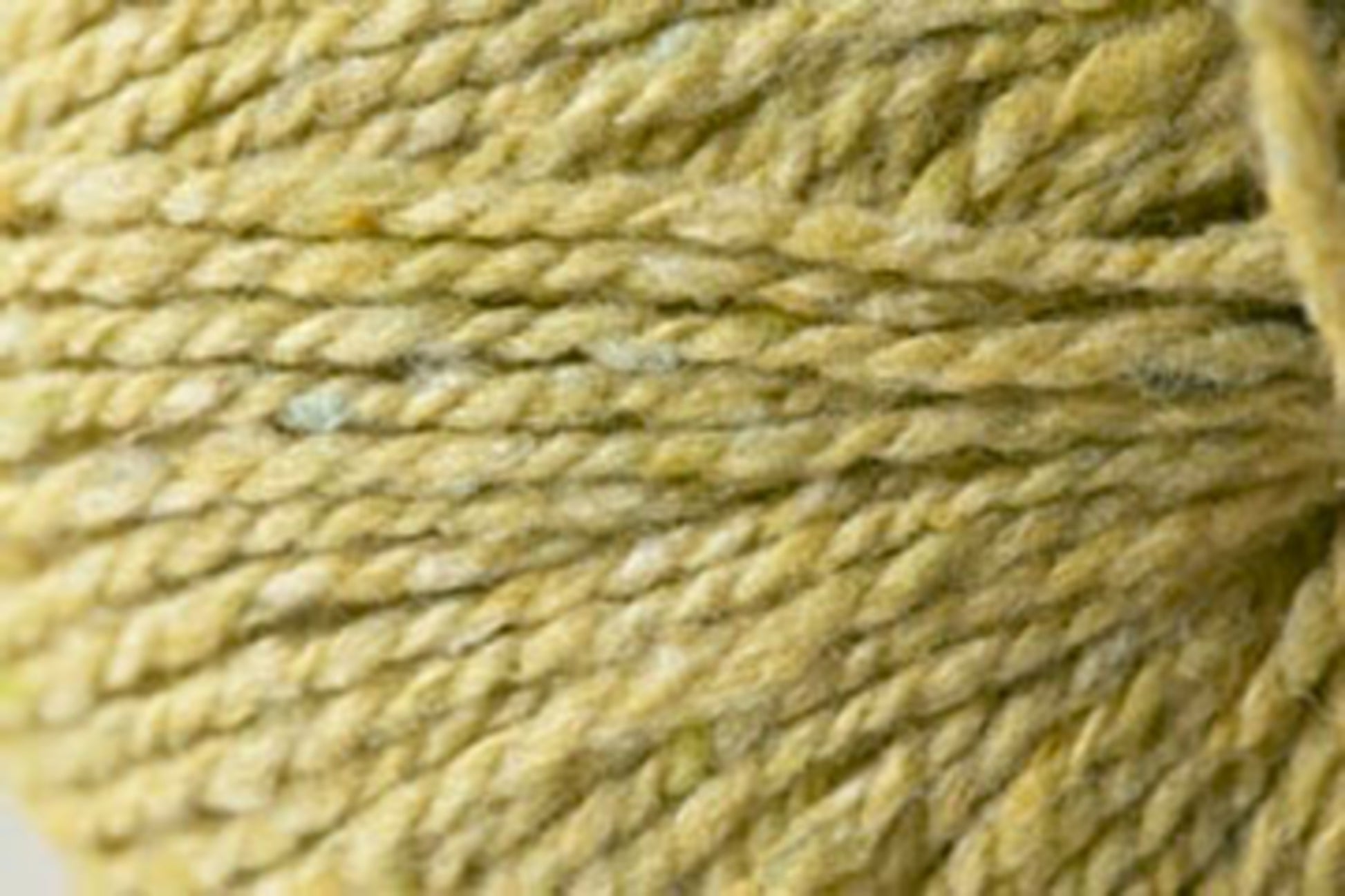 Close-up image of a ball of Jo Sharp Silkroad DK Tweed yarn by Kingfisher Yarn & Fibre in greenish-yellow. The photo showcases the texture and twist of this DK weight yarn, highlighting its slightly varied color tones and fibrous nature.