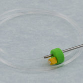 The Clover Circular Needle & Stitch Holder by Accessories Unlimited is a medical instrument that includes a long, clear flexible tube attached to a stainless steel needle with an adjustable cord length. The needle is partially covered by a green and yellow connector. This tool, reminiscent of circular needles, is laid out in a circular coil on a light grey surface.