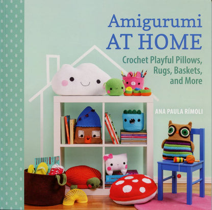 A book cover titled "Amigurumi At Home" by Ana Paula Rímoli, published by Martingale & Co, displays a shelf filled with vibrant crocheted home decor items. The cheerful designs include a smiling cloud, whimsical houses, an owl, and even a chair featuring a blue owl-shaped pillow.