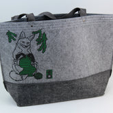 The Fox Felt Market Tote by Mum n Sun Ink from Bonnie Bishoff features a charming illustration of a fox knitting with green yarn on a gray polyester felt background. It includes two gray handles and is adorned with green pine branches above the fox, making it perfect for carrying your shawl pins and knit projects.