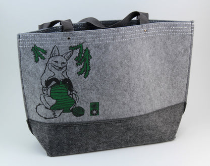 The Fox Felt Market Tote by Mum n Sun Ink from Bonnie Bishoff features a charming illustration of a fox knitting with green yarn on a gray polyester felt background. It includes two gray handles and is adorned with green pine branches above the fox, making it perfect for carrying your shawl pins and knit projects.